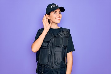 Young police woman wearing security bulletproof vest uniform over purple background smiling with hand over ear listening an hearing to rumor or gossip. Deafness concept.