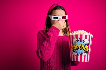 Young beautiful girl watching movie using 3d glasses eating box with popcorns smelling something...