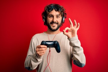 Young gamer man with curly hair and beard playing video game using joystick and headphones doing ok sign with fingers, excellent symbol