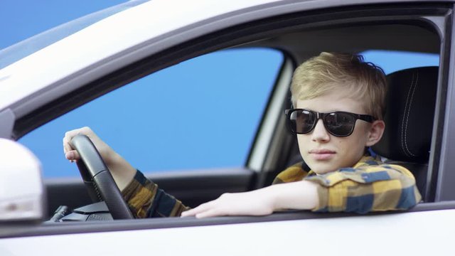 A Little Blonde Boy In Casual Clothes Is Travelling. Handsome Boy Is Driving Car With Greenscreen Background. He Is Sitting In The Car.