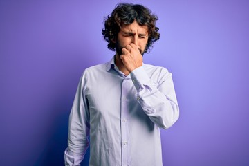 Young handsome business man with beard wearing shirt standing over purple background smelling something stinky and disgusting, intolerable smell, holding breath with fingers on nose. Bad smell