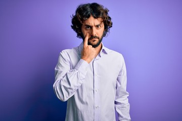 Young handsome business man with beard wearing shirt standing over purple background Pointing to the eye watching you gesture, suspicious expression