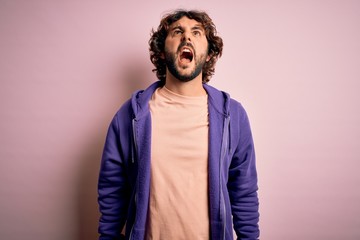 Obraz premium Young handsome sporty man with beard wearing casual sweatshirt over pink background angry and mad screaming frustrated and furious, shouting with anger. Rage and aggressive concept.