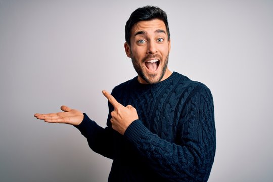 Young handsome man with beard wearing casual sweater standing over white background amazed and smiling to the camera while presenting with hand and pointing with finger.