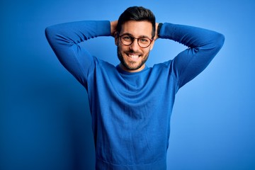 Young handsome man with beard wearing casual sweater and glasses over blue background relaxing and stretching, arms and hands behind head and neck smiling happy