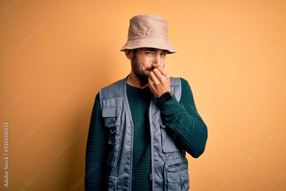 Canvas Prints Handsome tourist man with beard on vacation wearing explorer hat over yellow background smelling something stinky and disgusting, intolerable smell, holding breath with fingers on nose. Bad smell