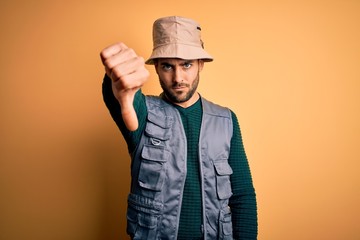 Handsome tourist man with beard on vacation wearing explorer hat over yellow background looking...