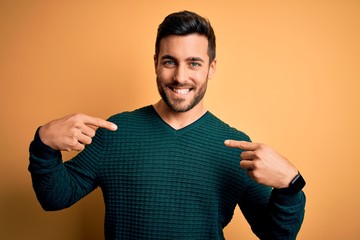 Young handsome man with beard wearing casual sweater standing over yellow background looking confident with smile on face, pointing oneself with fingers proud and happy.