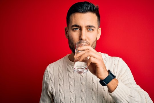 Young handsome man drinking glass of healthy water to refreshment standing over isolated red background