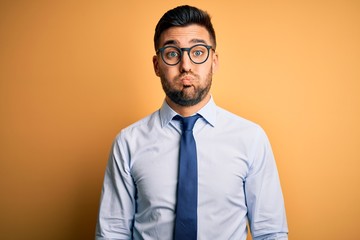 Young handsome businessman wearing tie and glasses standing over yellow background puffing cheeks...