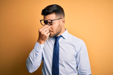 Young handsome businessman wearing tie and glasses standing over yellow background tired rubbing...