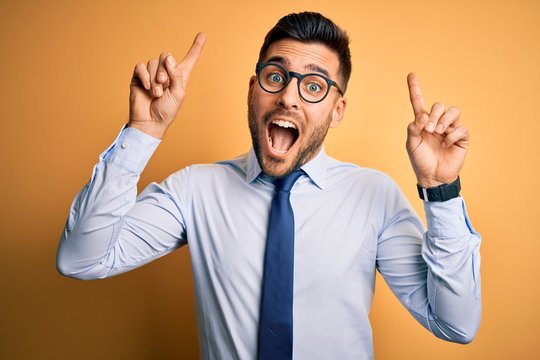 Young handsome businessman wearing tie and glasses standing over yellow background smiling amazed and surprised and pointing up with fingers and raised arms.