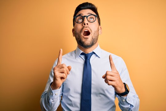 Young handsome businessman wearing tie and glasses standing over yellow background amazed and surprised looking up and pointing with fingers and raised arms.