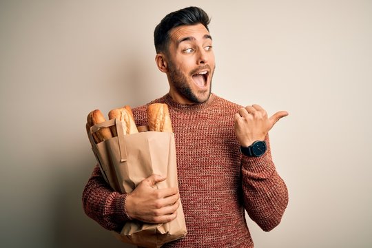 Young handsome man holding groceries paper bag of fresh baguette bread over isolated background pointing and showing with thumb up to the side with happy face smiling