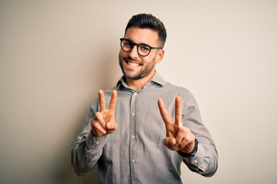 Young handsome man wearing elegant shirt and glasses over isolated white background smiling looking to the camera showing fingers doing victory sign. Number two.