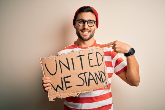 Handsome activist man protesting wearing glasses holding cardboard with unity message with surprise face pointing finger to himself