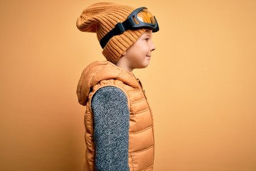 Young little caucasian kid wearing snow glasses and winter coat over yellow background looking to...