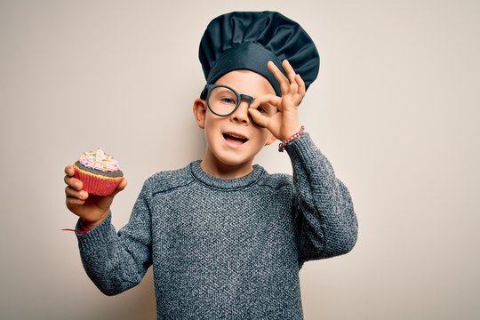 Young little caucasian cook kid wearing chef uniform and hat cooking cupcake with happy face smiling doing ok sign with hand on eye looking through fingers