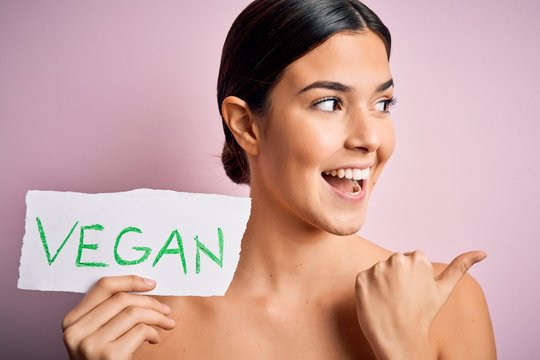 Young beautiful girl holding paper with vegan message over isolated pink background pointing and showing with thumb up to the side with happy face smiling