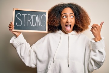 Young african american woman with afro hair holding blackboard with strong message pointing and showing with thumb up to the side with happy face smiling