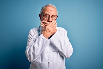 Fototapeta premium Senior grey haired doctor man wearing stethoscope and medical coat over blue background shocked covering mouth with hands for mistake. Secret concept.