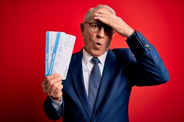 Senior grey haired business man holding airplane boarding pass over red background stressed with hand on head, shocked with shame and surprise face, angry and frustrated. Fear and upset for mistake.