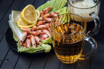 Boiled prawns on a black plate and a glass of beer on a wooden table. Shrimp with lemon on a lettuce leaf. Beer in a glass mug