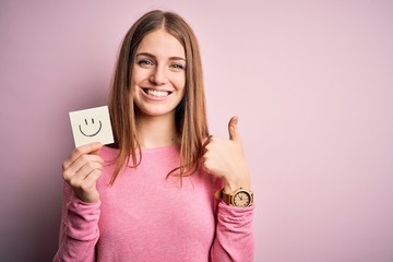 Young beautiful redhead woman holding reminder paper with smile emoji message happy with big smile doing ok sign, thumb up with fingers, excellent sign