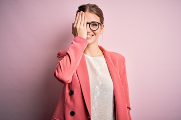 Young beautiful redhead woman wearing jacket and glasses over isolated pink background covering one eye with hand, confident smile on face and surprise emotion.