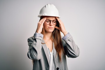Young beautiful redhead architect woman wearing security helmet over white background suffering from headache desperate and stressed because pain and migraine. Hands on head.
