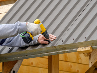 Men's hands in work gloves with a yellow screwdriver screw the roofing sheet to the roof of a...