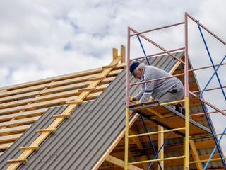 Naklejka premium Senior gray-haired Construction man using a screwdriver, fastens a roofing sheet to wooden rafters on the roof of a country house under construction. Physical activity of the seniors.