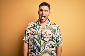 Young man with blue eyes on vacation wearing floral summer shirt over yellow background sticking tongue out happy with funny expression. Emotion concept.