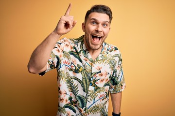 Young man with blue eyes on vacation wearing floral summer shirt over yellow background pointing...