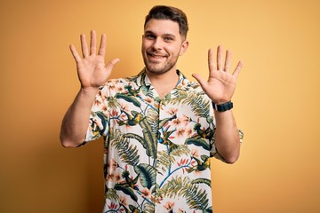 Young man with blue eyes on vacation wearing floral summer shirt over yellow background showing and pointing up with fingers number ten while smiling confident and happy.