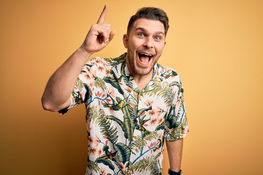 Young man with blue eyes on vacation wearing floral summer shirt over yellow background pointing finger up with successful idea. Exited and happy. Number one.