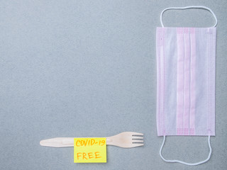 Wooden fork, pink medical mask and sticker with the inscription covid-19 free on a gray background. The concept of delivering safe, uninfected food during the quarantine coronavirus. Layout copy space