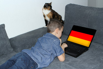 German flag on a laptop display, a little schoolboy in jeans lies on a sofa and scrolls, a cat sits...