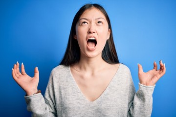 Young beautiful asian woman wearing casual sweater standing over blue isolated background crazy and mad shouting and yelling with aggressive expression and arms raised. Frustration concept.