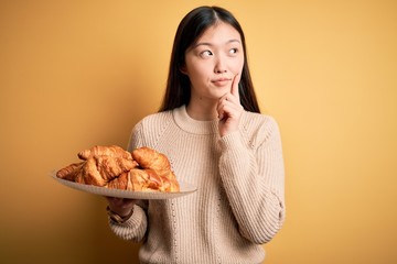 Young asian woman holding french pastry croissant over yellow isolated background serious face thinking about question, very confused idea