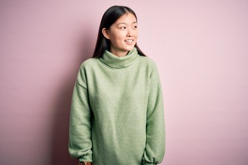 Young beautiful asian woman wearing green winter sweater over pink solated background looking away to side with smile on face, natural expression. Laughing confident.