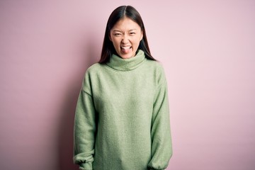 Young beautiful asian woman wearing green winter sweater over pink solated background sticking tongue out happy with funny expression. Emotion concept.