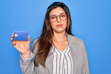 Young hispanic business woman holding credit card over blue isolated background with a confident expression on smart face thinking serious
