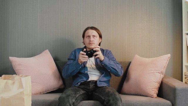 Young gamer holding joystick and playing video game sit alone on couch at home