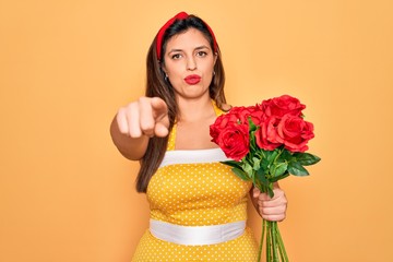 Young hispanic pin up woman wearing fashion sexy 50s style holding bouquet of red roses pointing with finger to the camera and to you, hand sign, positive and confident gesture from the front