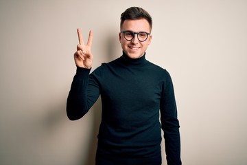 Young handsome caucasian man wearing glasses and casual sweater over isolated background showing and pointing up with fingers number two while smiling confident and happy.