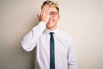 Young handsome caucasian business man wearing golden crown over isolated background covering one eye with hand, confident smile on face and surprise emotion.