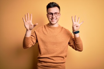 Young handsome caucasian man wearing glasses and casual winter sweater over yellow background showing and pointing up with fingers number nine while smiling confident and happy.