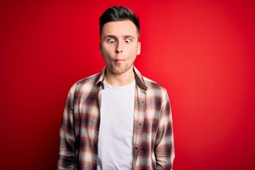 Obraz premium Young handsome caucasian man wearing casual modern shirt over red isolated background making fish face with lips, crazy and comical gesture. Funny expression.