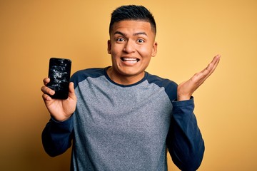 Young handsome latin man holding broken smartphone showing craked screen very happy and excited,...